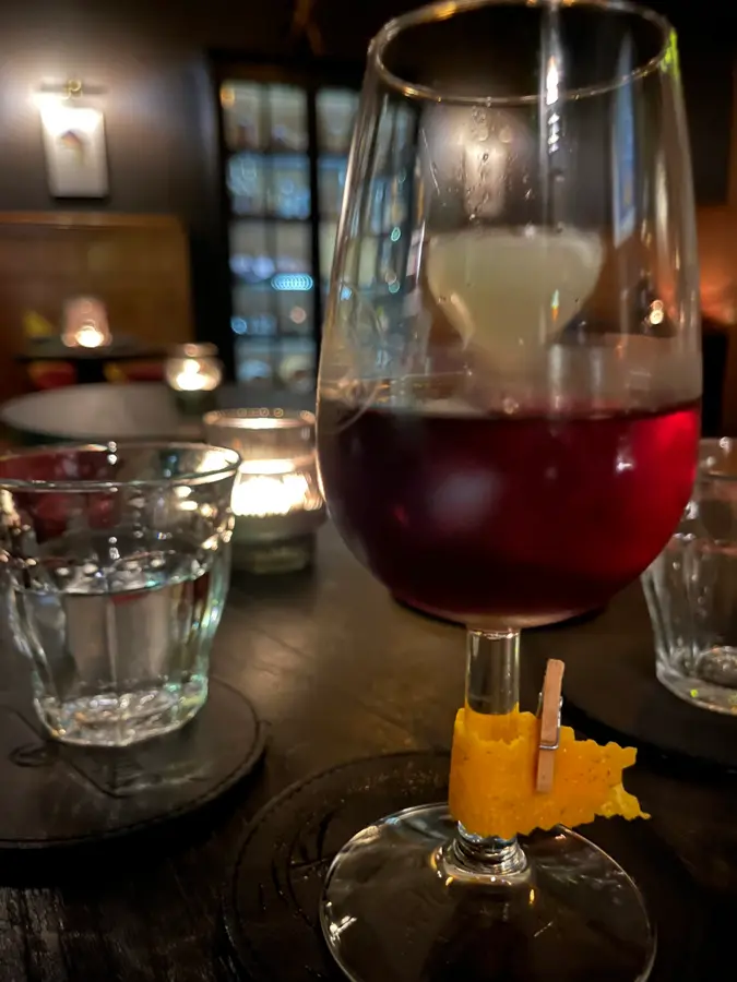 Tiefroter Drink im Stielglas mit Orangenzeste, im Hintergrund Kerzen und Sitznischen
